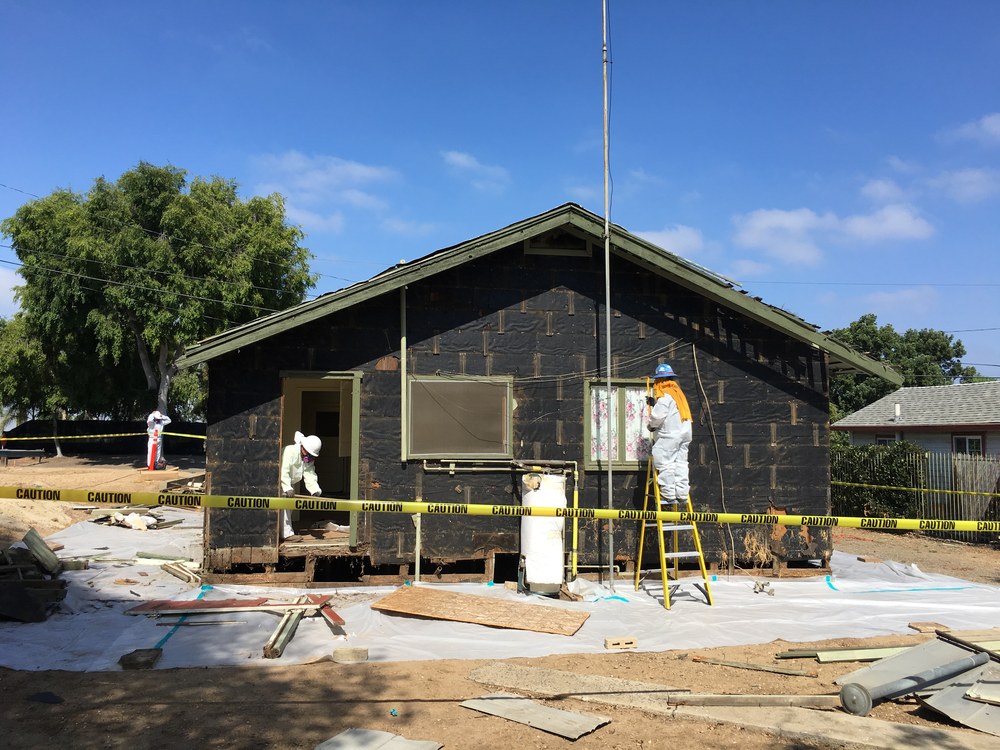 Workers in disposable coveralls removing exterior siding from a single-story house, with ground sheeting and caution tape marking the regulated work area