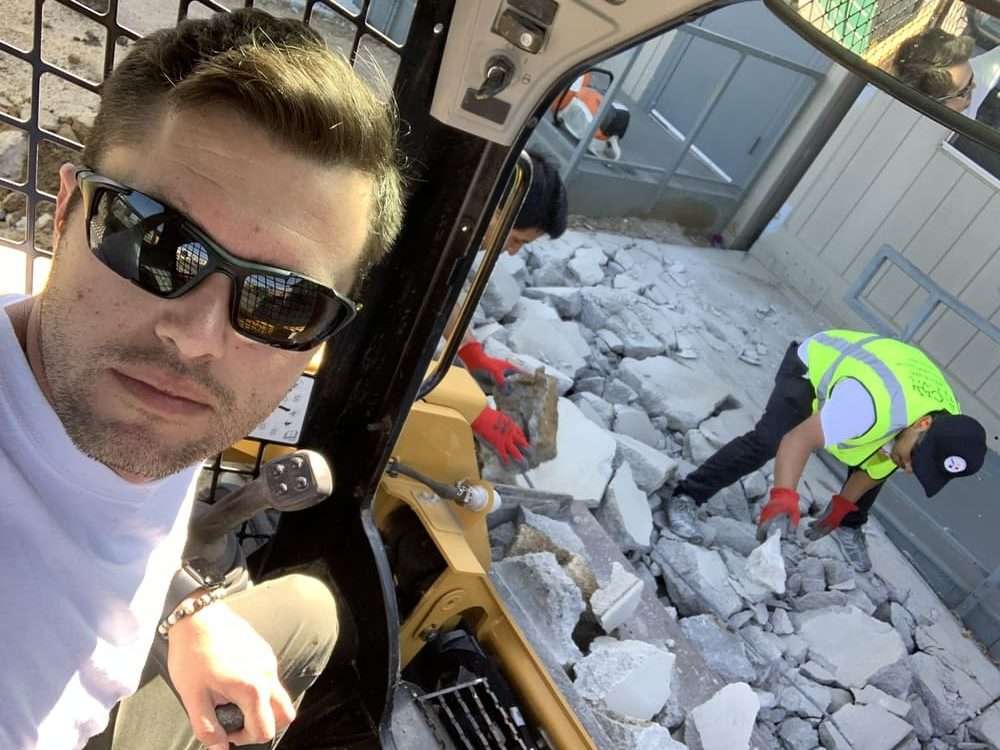 Skid-steer operator with workers handling concrete behind the machine.