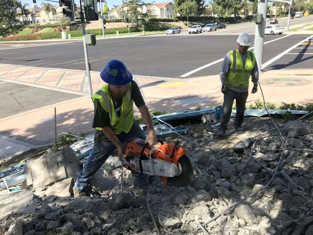 Two workers handling a cut-off saw among broken concrete near a public street