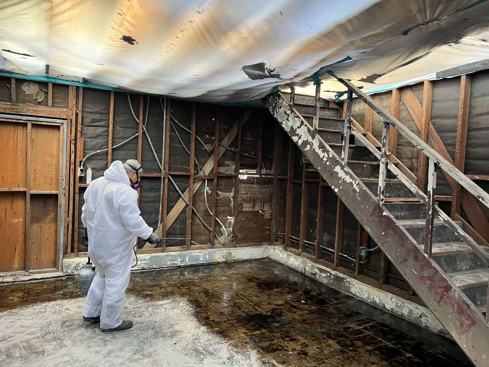 Person in protective clothing applying liquid inside a stripped interior with overhead sheeting