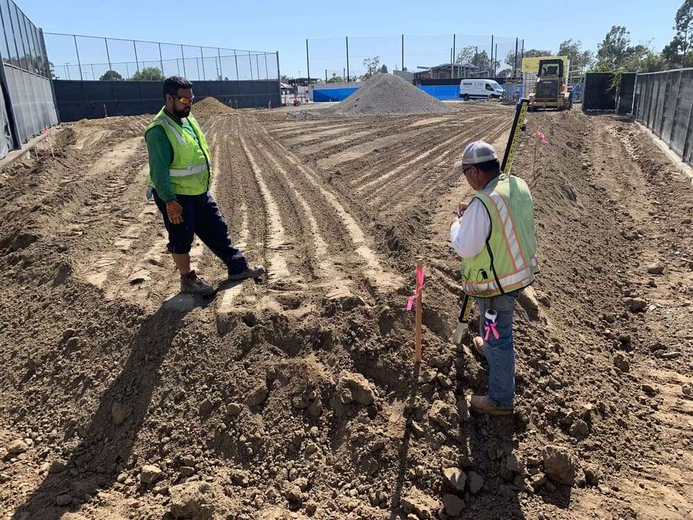 Two field personnel reviewing a measuring rod and marked stake within an active earthwork area
