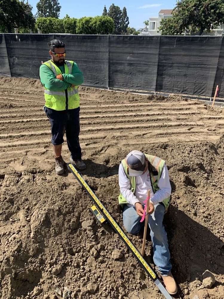 Field personnel checking a marked stake and measuring rod inside a selected excavation