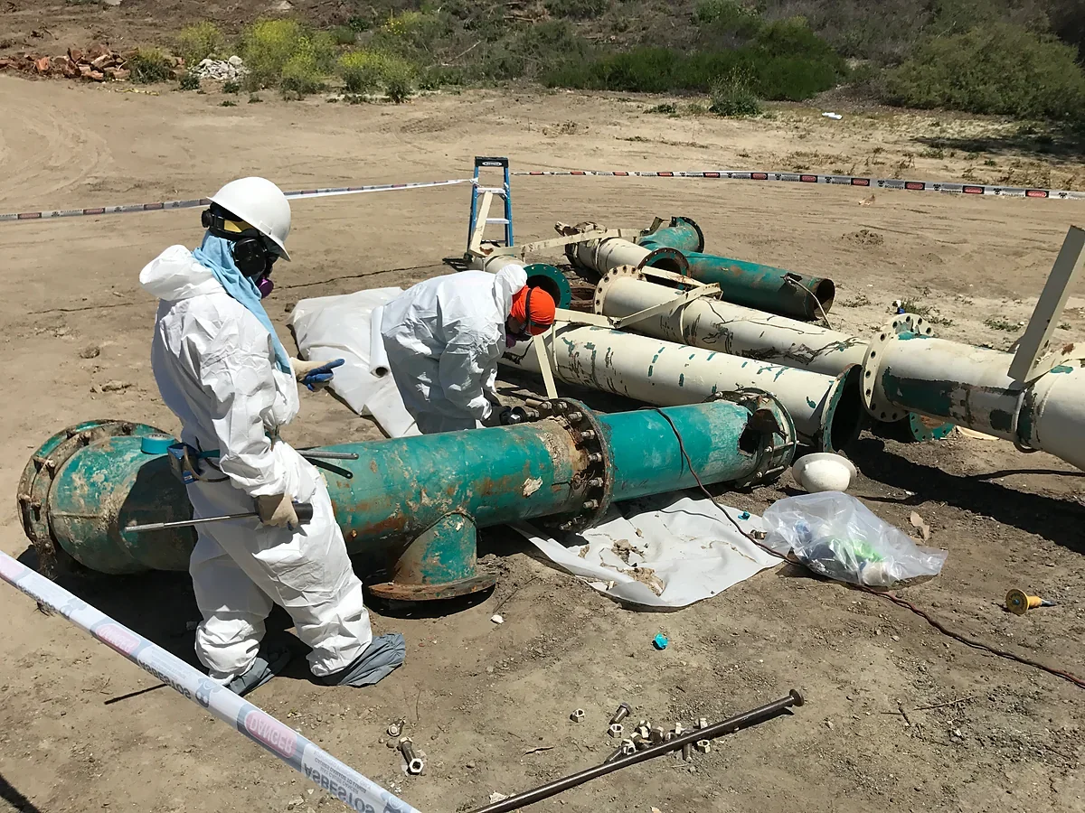 Two workers in protective clothing performing controlled work around removed industrial piping in a barricaded outdoor work area