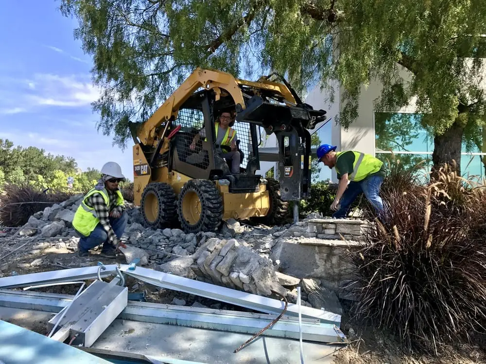 Three workers and a skid-steer loader with a hydraulic breaker removing concrete beside retained landscaping