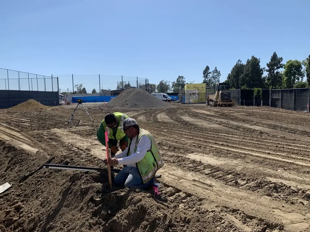 Two construction workers checking an issued field stake with survey equipment visible behind them