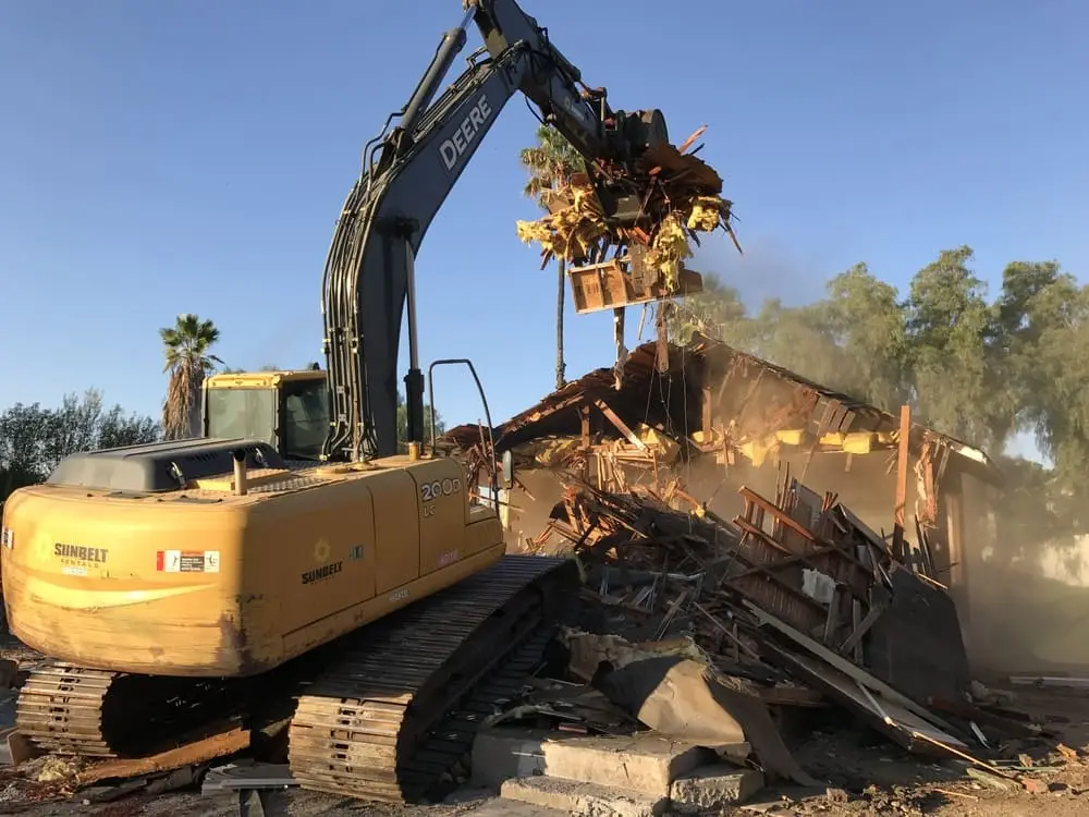 Tracked excavator lifting wood-framed demolition debris within a controlled work area