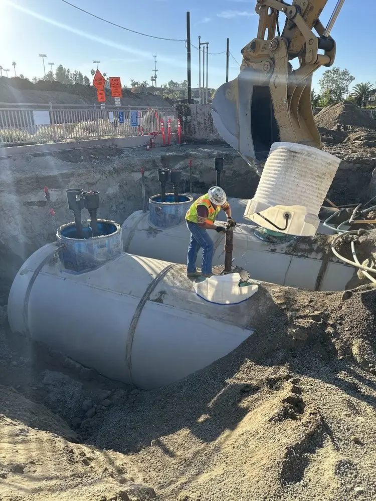 Worker standing on an exposed underground tank below an excavator bucket